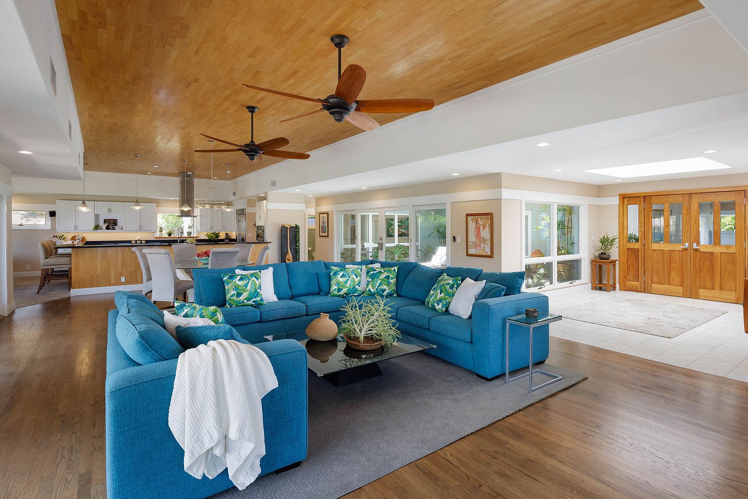 This is an interior shot of a spacious living room that seamlessly transitions into a modern kitchen. The living room features a large, blue sectional sofa with decorative pillows, a glass coffee table, and a gray area rug. The kitchen boasts a breakfast bar and modern appliances, creating an open and inviting atmosphere. The perspective is wide-angle, capturing the breadth of the combined living space.
