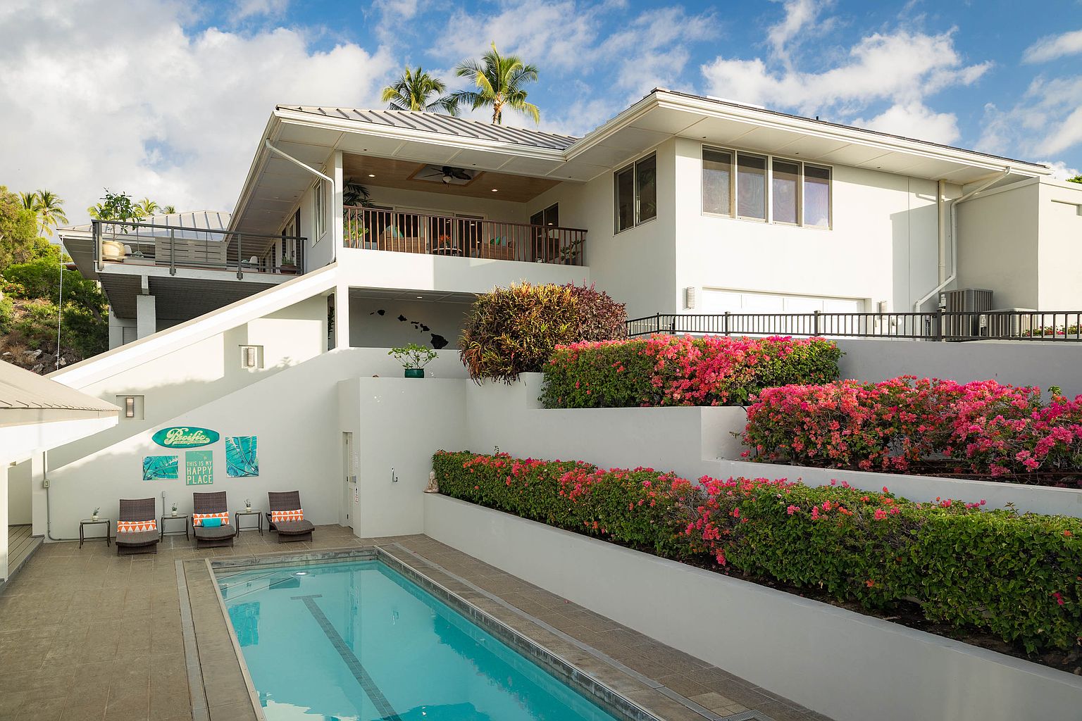 This image showcases a luxurious outdoor pool area of a modern home. The pool is surrounded by a tiled patio with lounge chairs, and tiered garden beds filled with vibrant pink flowers add color and texture. The white exterior of the house provides a clean backdrop, and the overall impression is one of relaxation and upscale living.