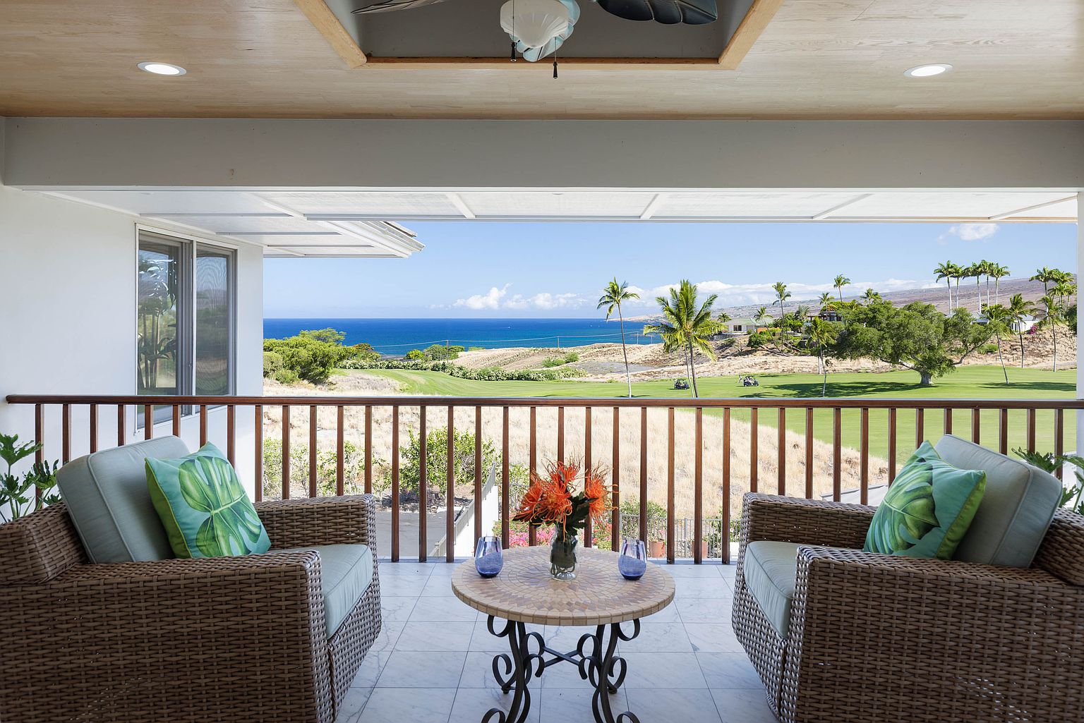 This image showcases a beautifully appointed balcony with wicker chairs and a small table adorned with flowers and blue glasses. The balcony overlooks a lush golf course and the ocean, providing a serene and luxurious outdoor living space. The view is framed by a wooden railing, enhancing the sense of privacy and exclusivity.