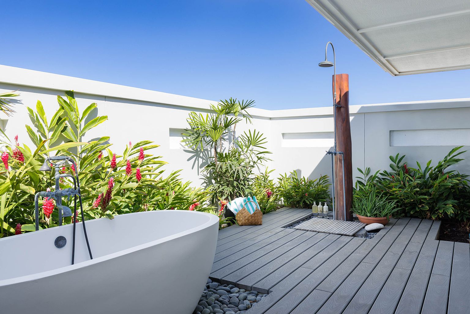 This image showcases a luxurious outdoor bathroom space, featuring a freestanding bathtub with vintage-style fixtures and an adjacent outdoor shower with a wooden post. The area is surrounded by lush tropical plants and a modern gray deck, creating a private and serene spa-like atmosphere. The design blends natural elements with contemporary architecture, offering a unique and upscale amenity.