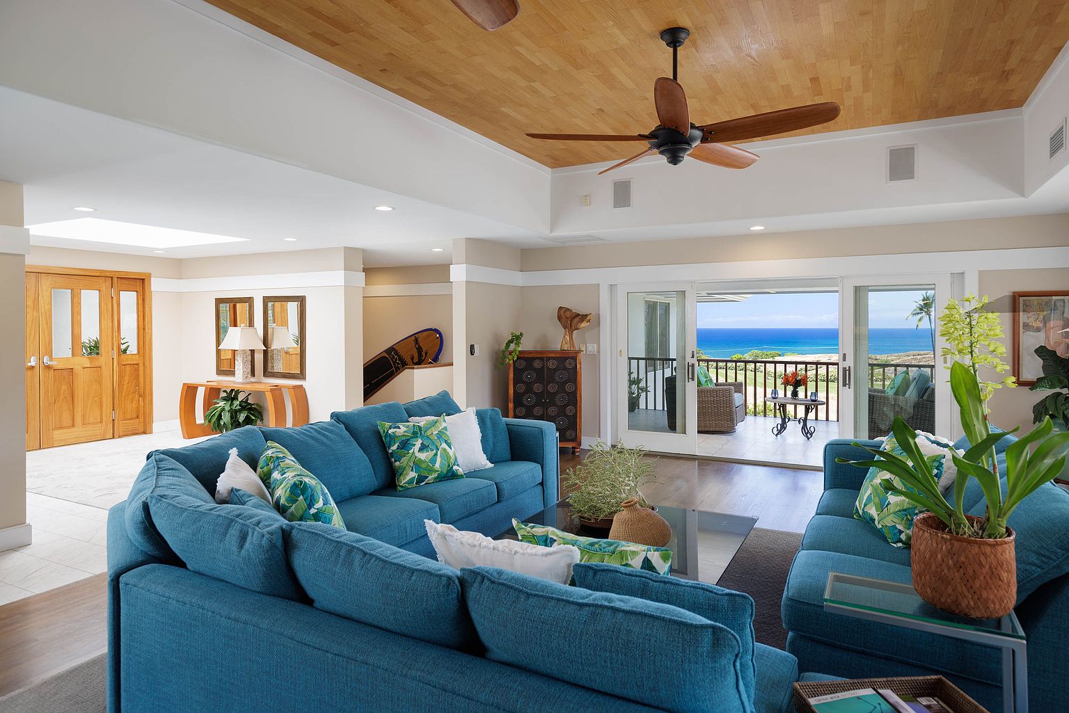 This is a bright and airy living room featuring a large, comfortable blue sectional sofa with decorative pillows. The room opens onto a balcony with ocean views, enhancing the indoor-outdoor living experience. The wooden ceiling and tasteful decor contribute to a relaxed, upscale atmosphere.