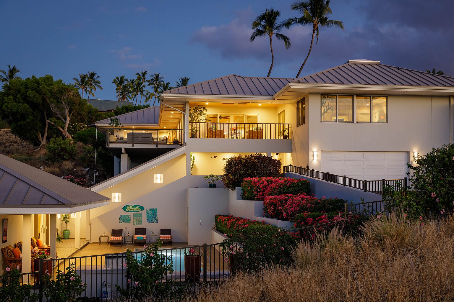 This image showcases the rear exterior of a multi-level home with a modern design. The property features multiple balconies, a swimming pool, and meticulously landscaped terraces with vibrant red flowers. The architecture is complemented by palm trees and a twilight sky, creating a luxurious and inviting atmosphere.