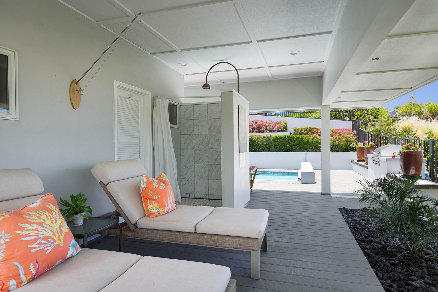 This image showcases a luxurious outdoor patio area, complete with comfortable lounge chairs adorned with vibrant pillows. The space features a stylish outdoor shower, a well-maintained deck, and a glimpse of a refreshing pool. Lush greenery and manicured landscaping add to the serene and upscale ambiance, perfect for relaxation and entertainment.