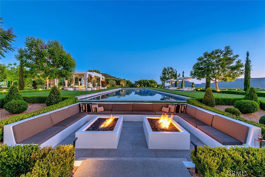 This image showcases a luxurious outdoor space featuring a modern seating area with built-in benches surrounding two fire pits. A long, rectangular pool stretches into the background, flanked by manicured lawns, mature trees, and a pergola. The scene exudes sophistication and relaxation, perfect for entertaining or enjoying quiet evenings.