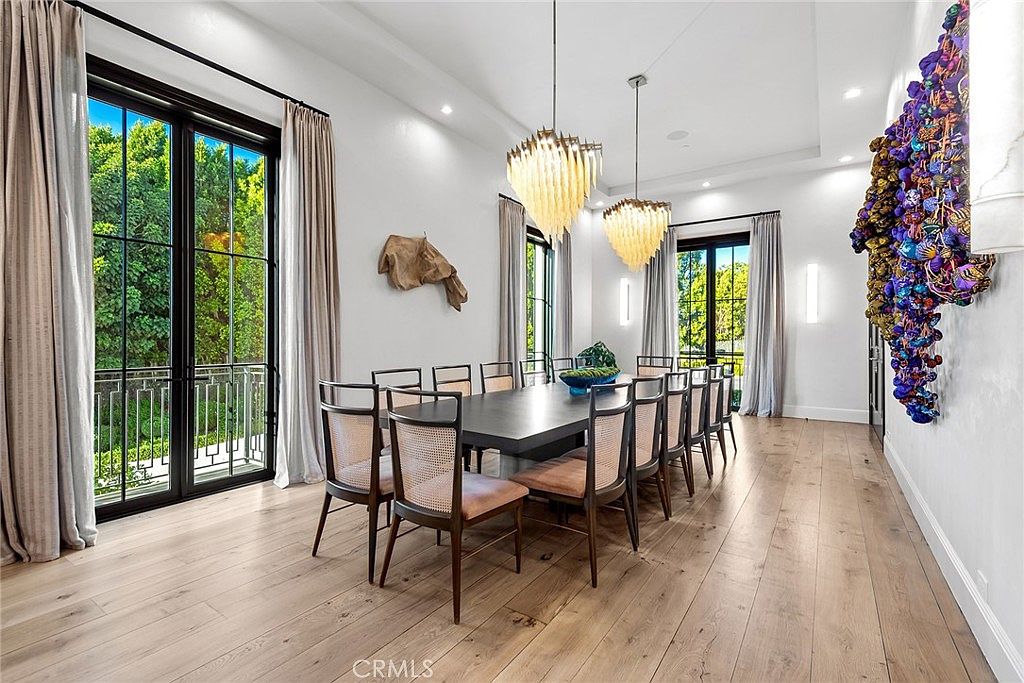 This is an interior shot of a dining room featuring a large, dark wood dining table surrounded by numerous chairs with woven backs and light-colored cushions. Two modern chandeliers hang above the table, and large windows with black frames and light-colored curtains provide natural light and a view of the greenery outside. The room has light wood flooring and white walls, creating a bright and elegant atmosphere.