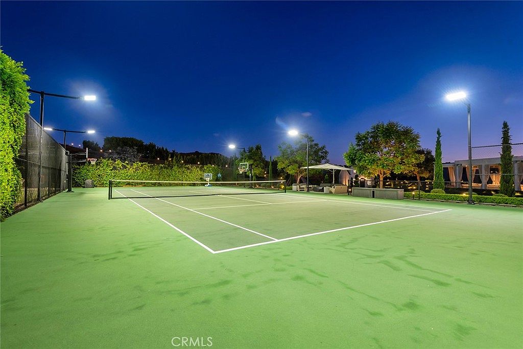 The image showcases a well-lit tennis court at night, featuring a vibrant green surface and clearly marked lines. A net divides the court, and lighting fixtures illuminate the playing area. In the background, there are trees and a glimpse of a building, suggesting a luxurious residential setting.
