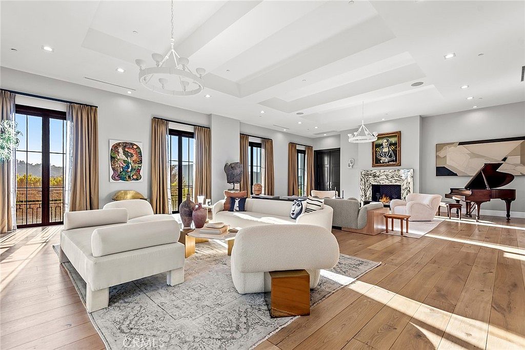 This is a spacious and luxurious living room featuring high ceilings with recessed lighting and decorative chandeliers. The room is furnished with modern, plush seating arrangements, including sofas and armchairs, arranged around a central coffee table on a patterned rug. A grand piano sits near a fireplace, adding to the room's elegance and sophistication, while large windows with curtains provide natural light and views.
