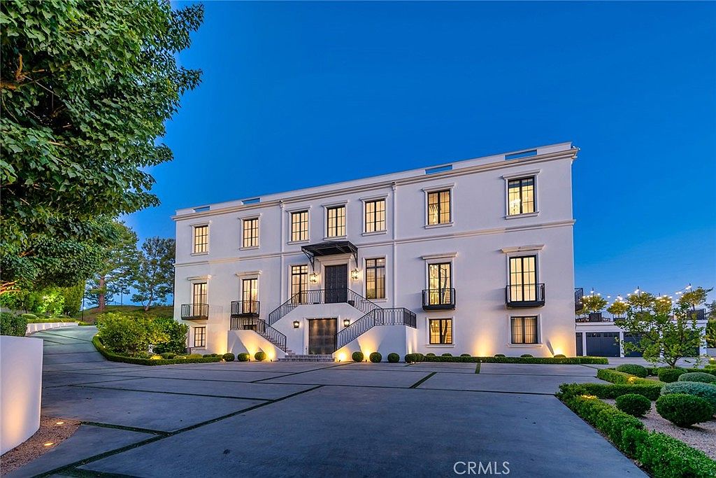 This is a front exterior view of a grand, three-story white mansion. The building features symmetrical window placement, black framed windows, and small balconies. A wide driveway leads up to the entrance, which is accessed by a set of stairs with black railings, and the landscaping includes manicured hedges and trees.