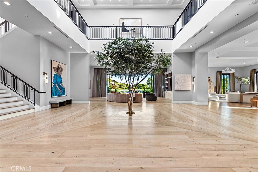 This grand foyer features light hardwood flooring in a herringbone pattern, a striking central tree, and a second-story balcony with black iron railings. A staircase with matching railings is visible on the left, and artwork adorns the walls, creating an elegant and inviting entrance. The space is bright and airy, emphasizing the home's luxurious design.