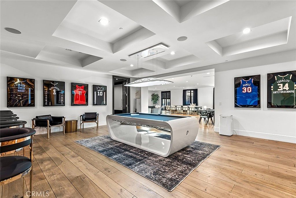 This interior shot showcases a modern living room with a unique pool table as a centerpiece, set upon a patterned rug. The room features hardwood flooring, recessed lighting, and a series of framed sports jerseys adorning the wall, adding a personalized touch. The space is open and well-lit, suggesting a comfortable and stylish entertainment area.
