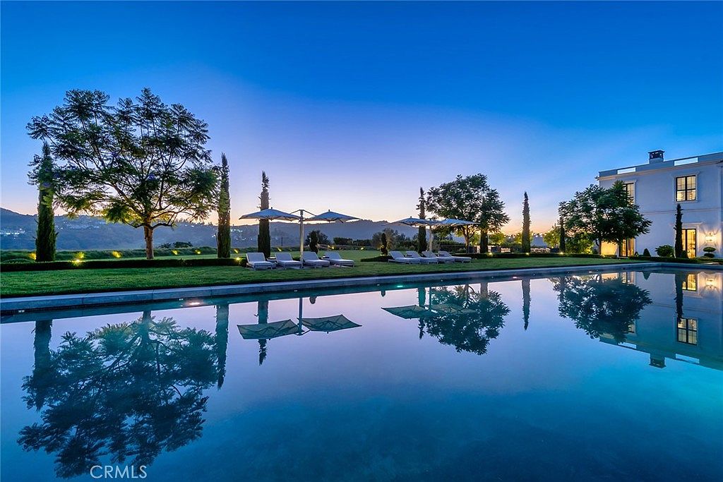 This image showcases a luxurious pool area at dusk, reflecting the surrounding landscape and architecture. The pool's still surface mirrors the trees, umbrellas, and the elegant white building in the background, creating a serene and upscale ambiance. Lounge chairs are neatly arranged on the manicured lawn, suggesting a relaxing and private retreat.