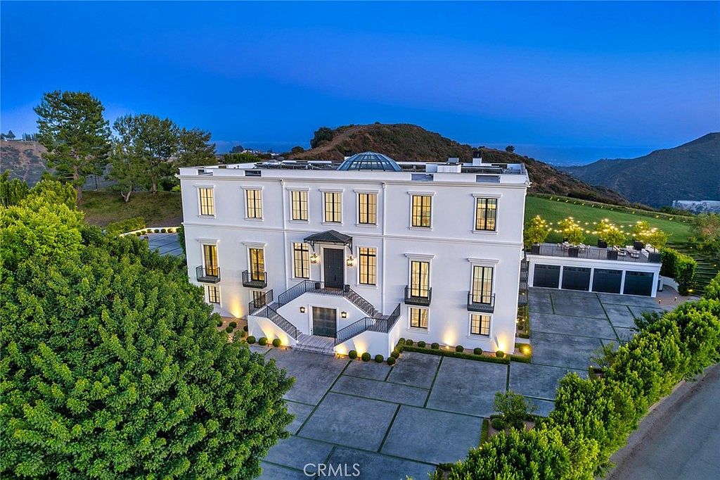 This is a stunning front view of a grand, three-story white mansion. The property features a symmetrical facade with black-framed windows, small balconies, and a prominent entryway with stairs leading up to the front door. A well-manicured lawn and mature trees frame the house, adding to its curb appeal and sense of luxury.