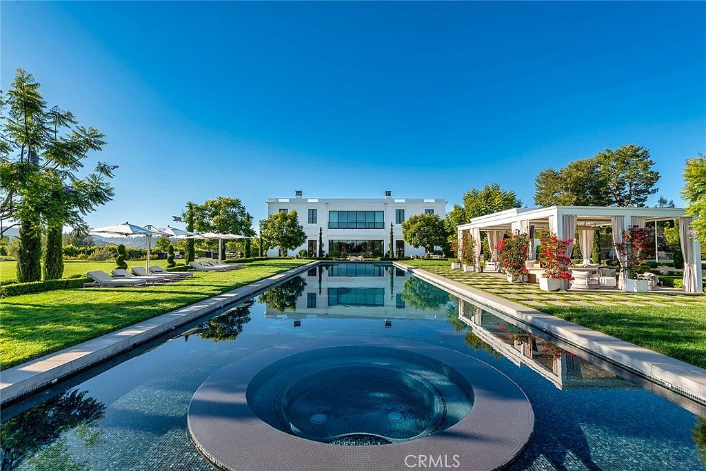 This image showcases a luxurious backyard featuring a long, rectangular pool with an integrated circular spa. The pool reflects the grand white house in the background, surrounded by manicured lawns, mature trees, and a covered outdoor lounge area. The scene exudes elegance and relaxation, perfect for high-end real estate.