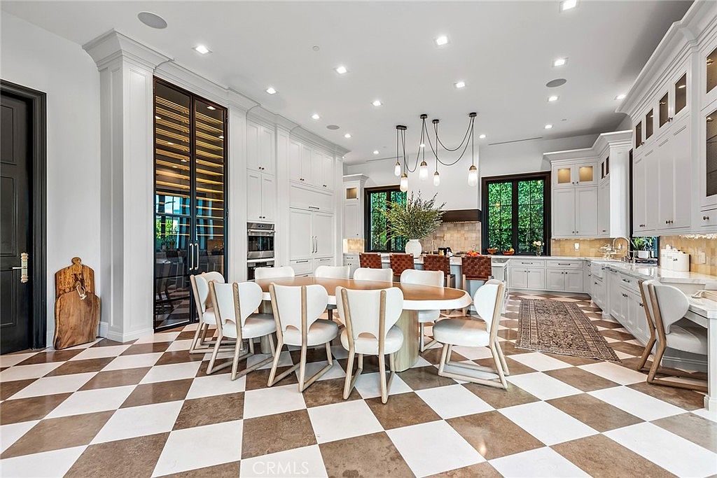 This is a spacious kitchen and dining area featuring a checkered tile floor and a large oval dining table surrounded by modern chairs. The kitchen boasts white cabinetry, stainless steel appliances, and a central island with pendant lighting. Large windows provide natural light, enhancing the bright and airy atmosphere of the space.