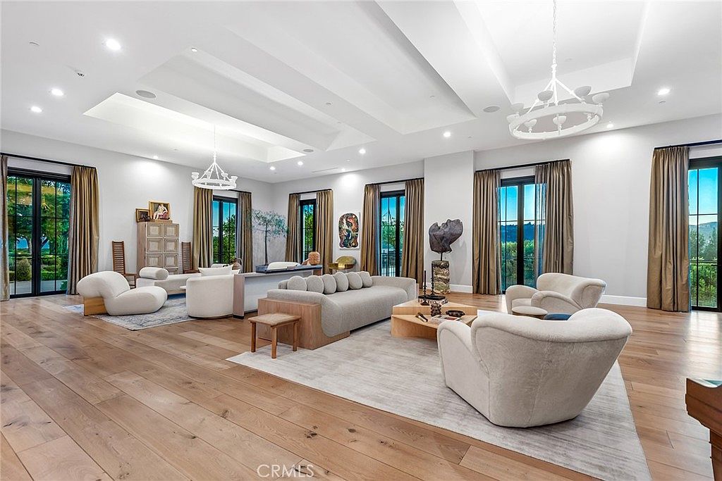 This is a spacious living room featuring hardwood floors, a neutral color palette, and abundant natural light from large windows. The room is furnished with a contemporary sofa, armchairs, and a stylish coffee table, creating a comfortable and inviting atmosphere. The chandeliers and artwork add a touch of elegance to the space.