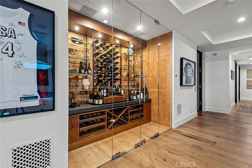 This interior shot showcases a sophisticated wine cellar with a glass enclosure, wooden shelving, and a dark countertop. The cellar is well-stocked with wine bottles and glassware, creating an inviting space for wine enthusiasts. The hardwood flooring and adjacent hallway add to the luxurious feel of the home.