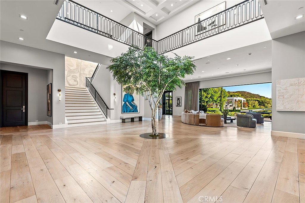 This grand foyer features a soaring two-story ceiling, a striking staircase with black iron railings, and light wood flooring laid in a herringbone pattern. A large tree is centered in the space, adding a natural element, while artwork and comfortable seating are visible in the adjacent living area. The overall impression is one of luxury and spaciousness.