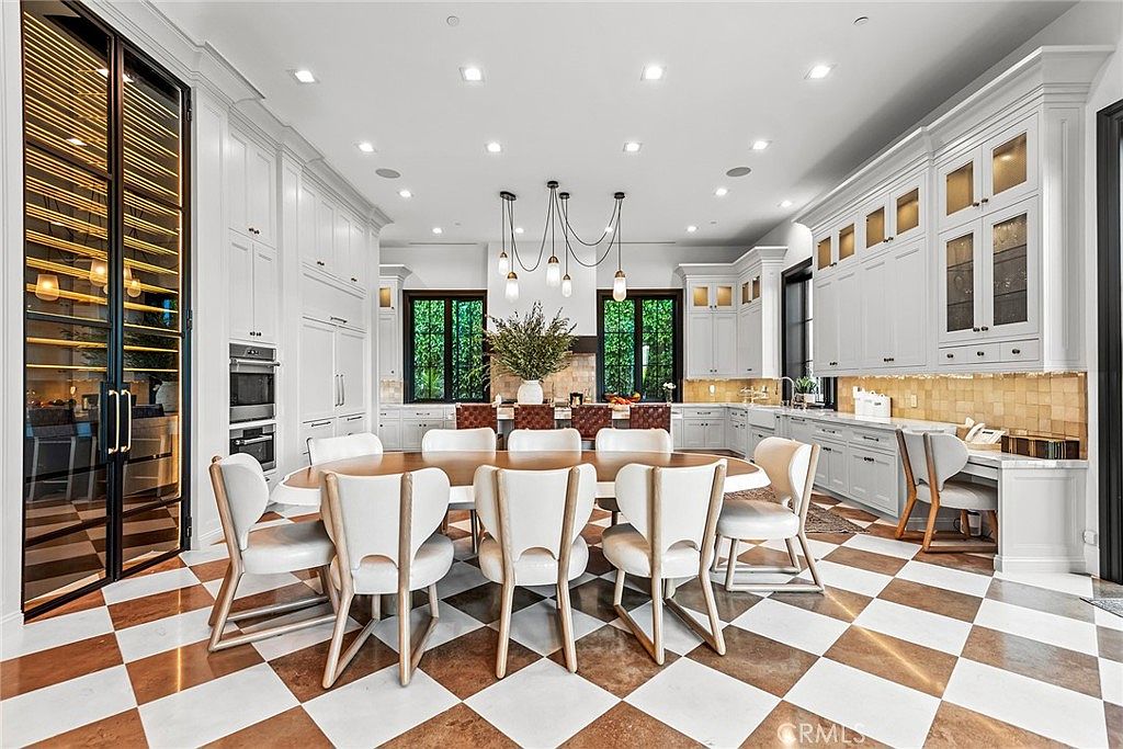 This is a spacious and luxurious kitchen featuring a large dining table surrounded by elegant chairs, perfect for family meals or entertaining guests. The kitchen boasts custom white cabinetry, high-end appliances, and a striking checkered floor pattern, creating a sophisticated and inviting atmosphere. Natural light floods the space through large windows, enhancing the overall brightness and appeal.