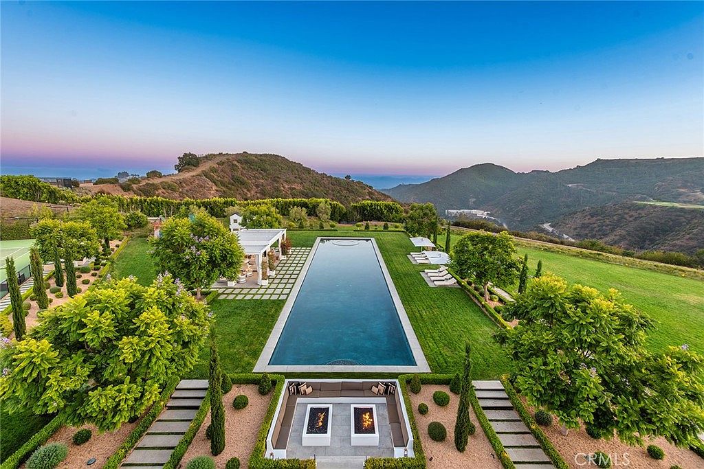 This aerial view showcases a luxurious estate with meticulously landscaped grounds. A long, rectangular pool is the focal point, flanked by lush green lawns, manicured hedges, and mature trees. A modern outdoor lounge area with fire pits sits at one end of the pool, while a covered patio area is located at the other end. The property is nestled against a backdrop of rolling hills and mountains, offering stunning views and a sense of privacy.