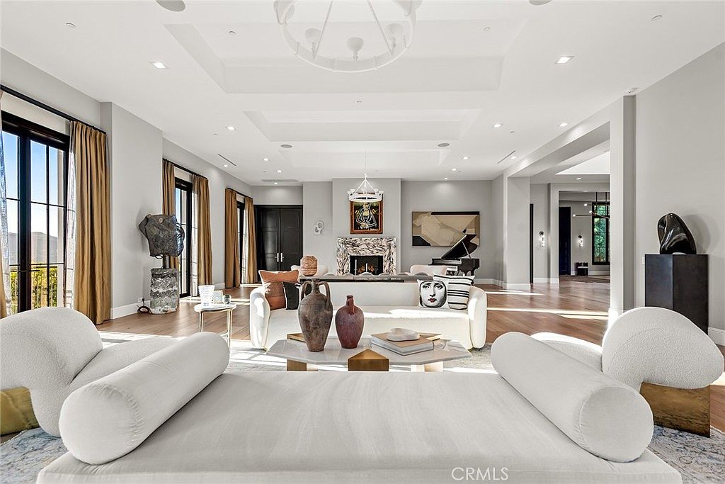 This is a spacious and luxurious living room featuring a neutral color palette with hardwood floors and recessed lighting. The room is furnished with modern white sofas, a marble coffee table, and various decorative art pieces, including sculptures and paintings. Large windows with elegant curtains provide natural light and a view to the outside, creating an inviting and sophisticated atmosphere.