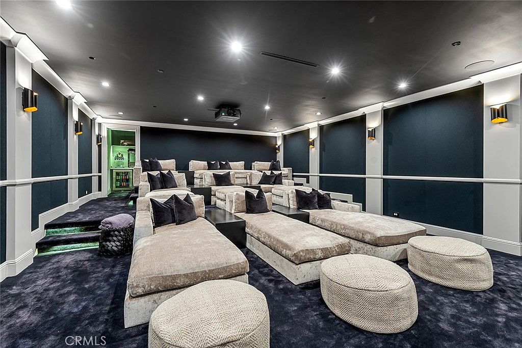 This is a luxurious home theater featuring tiered seating with plush chaise lounges and ottomans, creating a comfortable and immersive viewing experience. The room is decorated with dark blue and white striped walls, sconce lighting, and a dark ceiling, enhancing the cinematic atmosphere. A projector is mounted on the ceiling, ready for entertainment.
