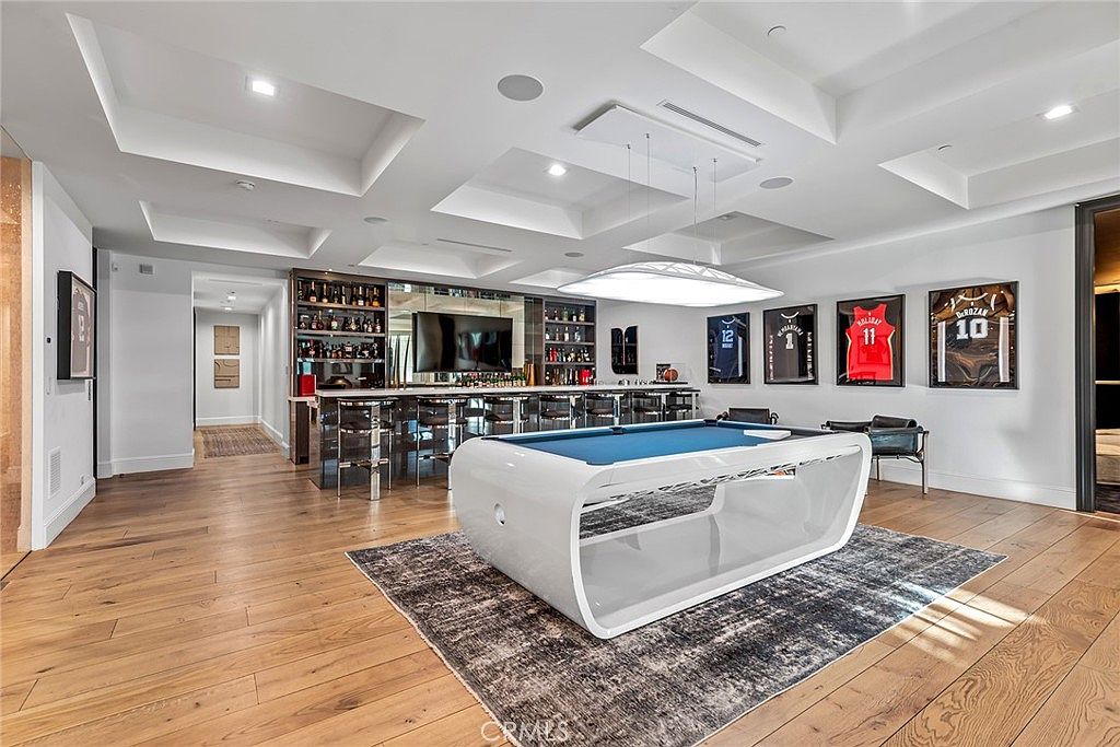This interior shot showcases a luxurious living room featuring a modern pool table on a patterned rug, a fully stocked bar with mirrored backsplash, and framed sports jerseys adorning the wall. The room is illuminated by unique ceiling lighting and features hardwood flooring, creating a sophisticated and entertainment-ready space.