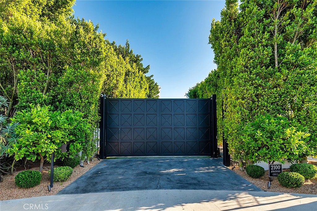 The image showcases an impressive gated entryway to a property, featuring a large, dark-colored gate with a geometric design, flanked by lush green hedges and trees. A well-maintained driveway leads up to the gate, and a small sign displaying the address is visible on the right. The scene conveys a sense of privacy, security, and established landscaping, suggesting a high-end property.