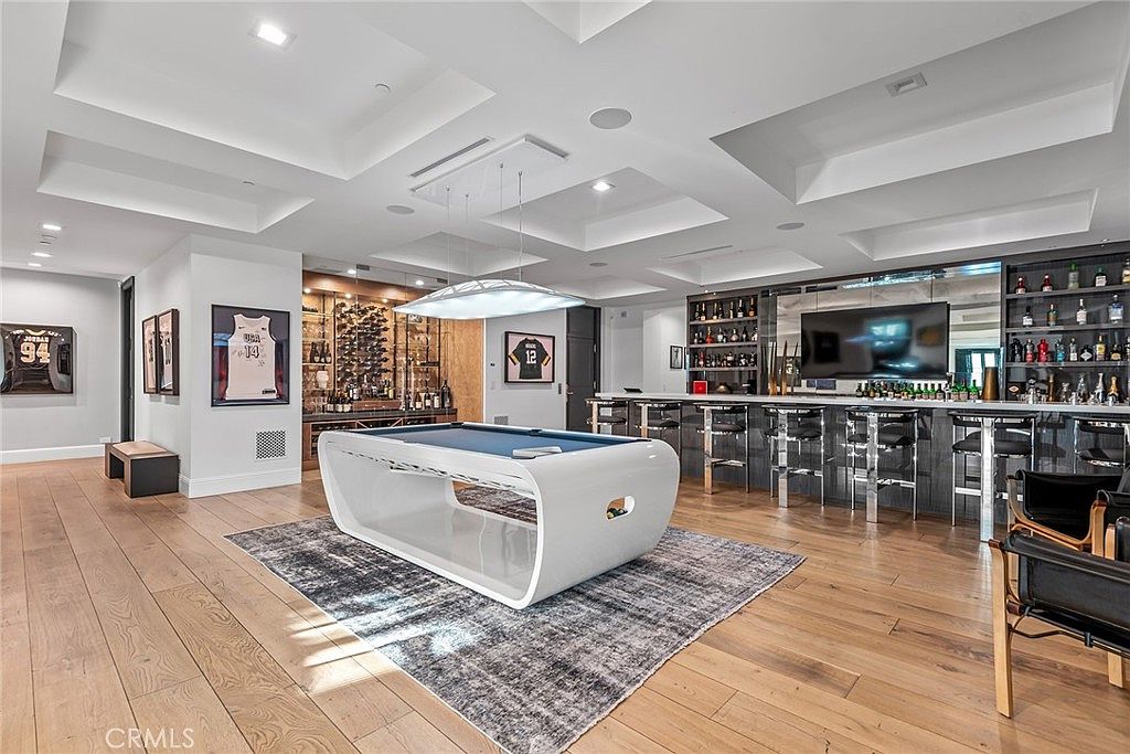 This interior shot showcases a luxurious living room featuring a modern pool table on a patterned rug, complemented by a well-stocked bar area with stylish seating. The room is adorned with sports memorabilia and features elegant lighting fixtures, creating a sophisticated and entertaining space. The hardwood flooring adds warmth and richness to the overall design.