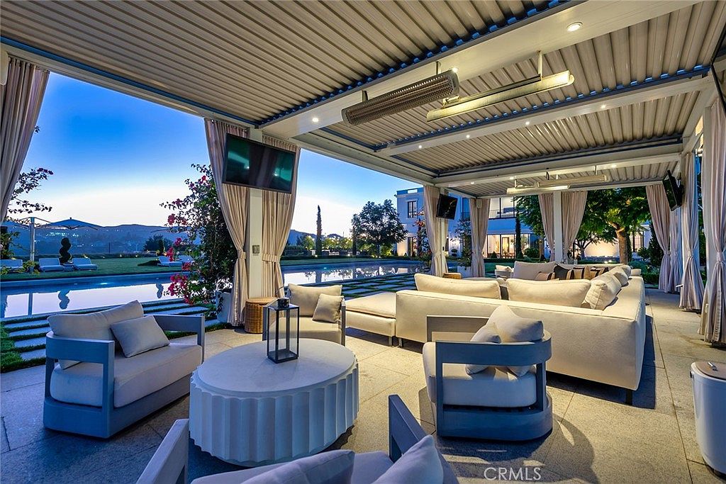 This image showcases a luxurious outdoor patio area, featuring comfortable seating arrangements under a covered structure with elegant curtains. A swimming pool is visible in the background, adding to the upscale ambiance. The space is designed for relaxation and entertainment, with a focus on comfort and style.