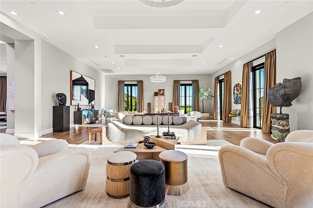 This is a spacious and luxurious living room featuring high ceilings, hardwood floors, and large windows with elegant drapery. The room is furnished with modern, comfortable seating, including a curved sofa and plush armchairs, arranged around a stylish coffee table. Artwork and decorative objects add to the sophisticated ambiance, creating an inviting and upscale living space.