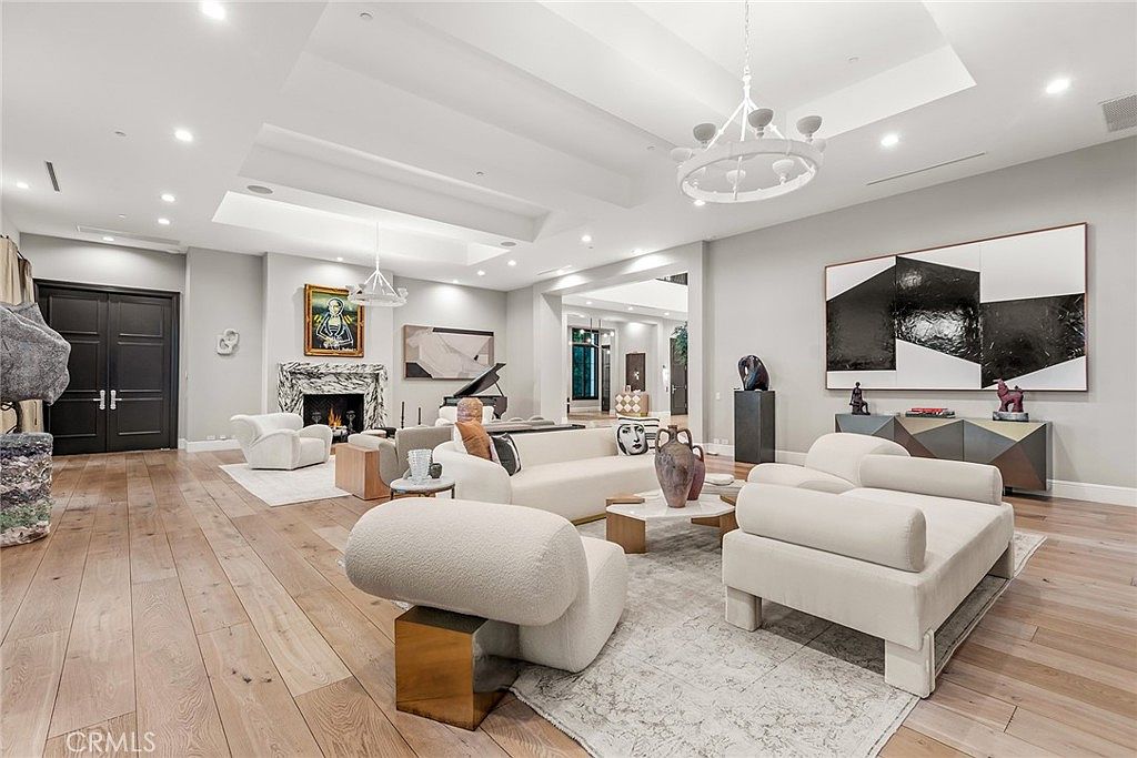 This is a spacious and luxurious living room featuring hardwood floors, a large area rug, and modern furniture. The room is well-lit with recessed lighting and a statement chandelier. Artwork and sculptures add to the sophisticated ambiance, creating an inviting and stylish space.