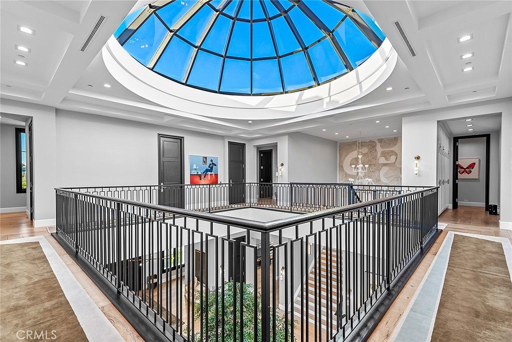 This interior shot showcases a luxurious hallway with a striking architectural feature: a large, domed skylight that floods the space with natural light. The hallway features a black metal railing overlooking a lower level with lush greenery, adding depth and visual interest. The flooring is hardwood, complemented by area rugs, and the walls are painted in a neutral tone, creating a bright and airy atmosphere.