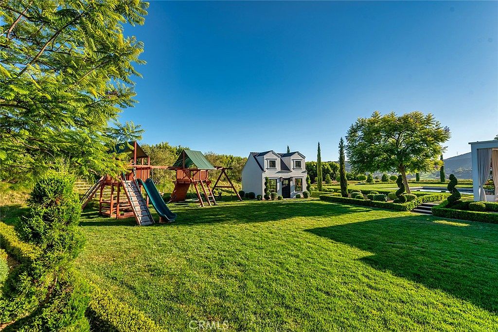 This image showcases a beautifully landscaped yard with lush green grass, manicured hedges, and mature trees under a clear blue sky. A charming playhouse and playground set are visible, adding a touch of family-friendly appeal. The overall impression is one of a well-maintained and inviting outdoor space, perfect for relaxation and recreation.