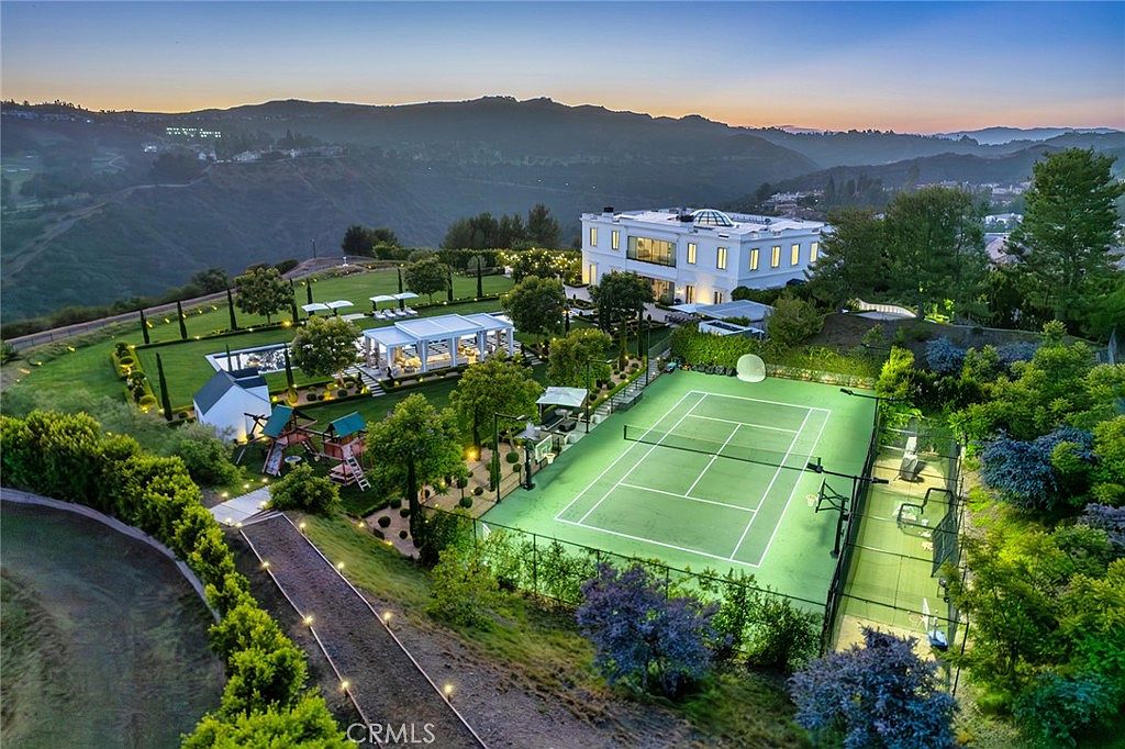 This aerial view showcases a luxurious estate nestled in a hilly landscape. The property features a grand white mansion, a tennis court, a swimming pool with a cabana, and meticulously manicured lawns and gardens. A winding pathway leads through the grounds, highlighting the expansive and private nature of the estate.