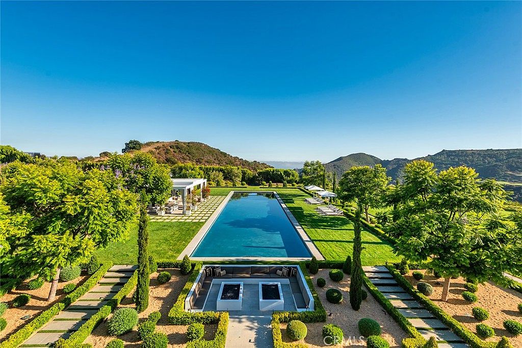 This is an aerial view of a luxurious backyard featuring a long, rectangular swimming pool as the focal point. The pool is surrounded by manicured lawns, formal gardens with trimmed hedges, and mature trees. A sunken seating area with a fire pit is located at one end, while lounge chairs and a covered patio are positioned at the other, creating an inviting and upscale outdoor living space.