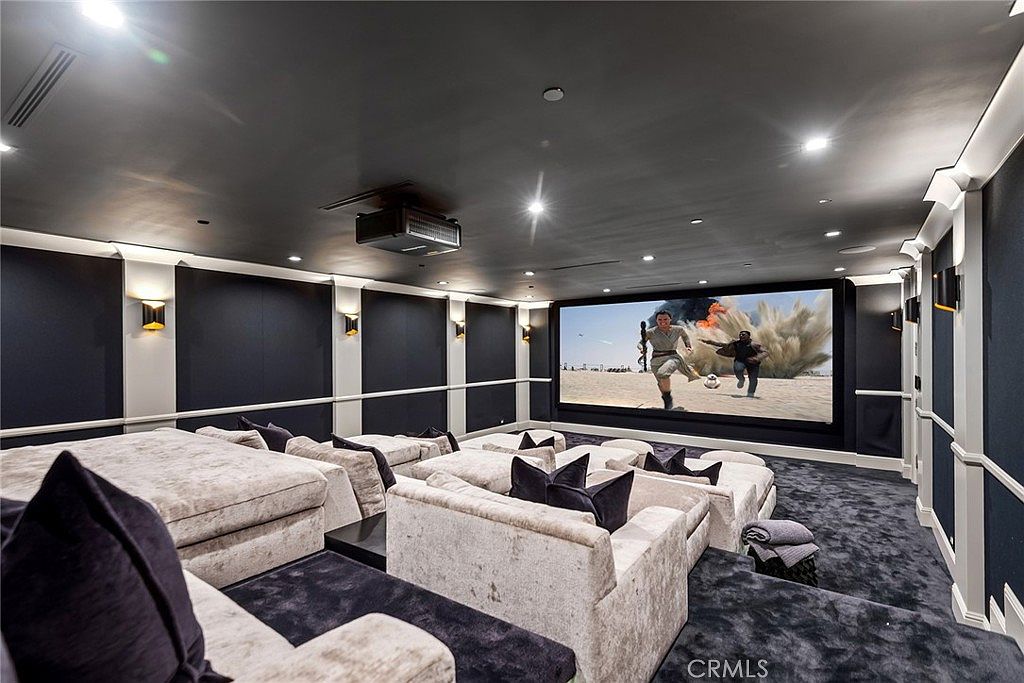 This is a luxurious home theater room featuring tiered seating with plush, modular sofas and dark throw pillows. A large screen displays an action scene, and the room is equipped with a projector mounted on the dark ceiling. The walls are paneled with dark fabric and white trim, creating a sophisticated and immersive viewing experience.