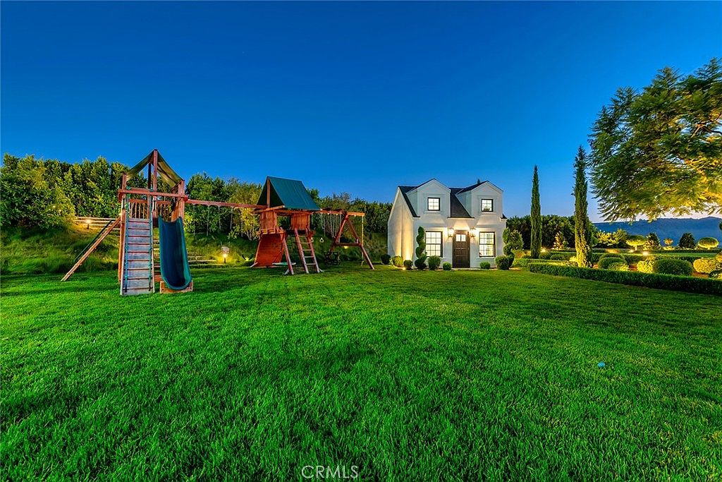 This image showcases a beautifully landscaped yard with a well-maintained lawn, a charming white house, and a wooden playset. The yard features manicured hedges, mature trees, and decorative lighting, creating an inviting and picturesque outdoor space. The scene is captured at dusk, highlighting the property's serene and elegant ambiance.