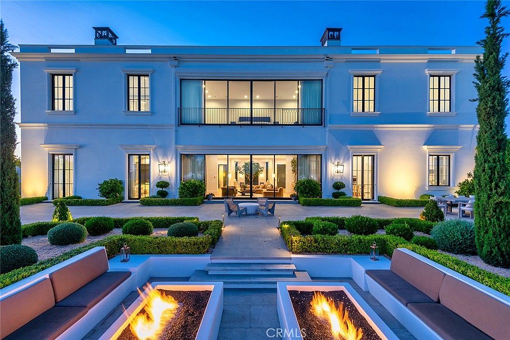 This is a rear exterior view of a luxurious two-story home featuring a symmetrical design with multiple windows and doors. The property boasts meticulously manicured landscaping, including trimmed hedges and tall trees. In the foreground, there are modern fire pits with built-in seating, creating an inviting outdoor living space.