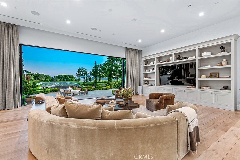 This is a luxurious living room featuring a large, curved sofa and a custom built-in entertainment center. The room opens to an expansive outdoor area with a pool and manicured garden through a large sliding glass door, creating a seamless indoor-outdoor living experience. The hardwood floors and neutral color palette contribute to a sophisticated and inviting atmosphere.