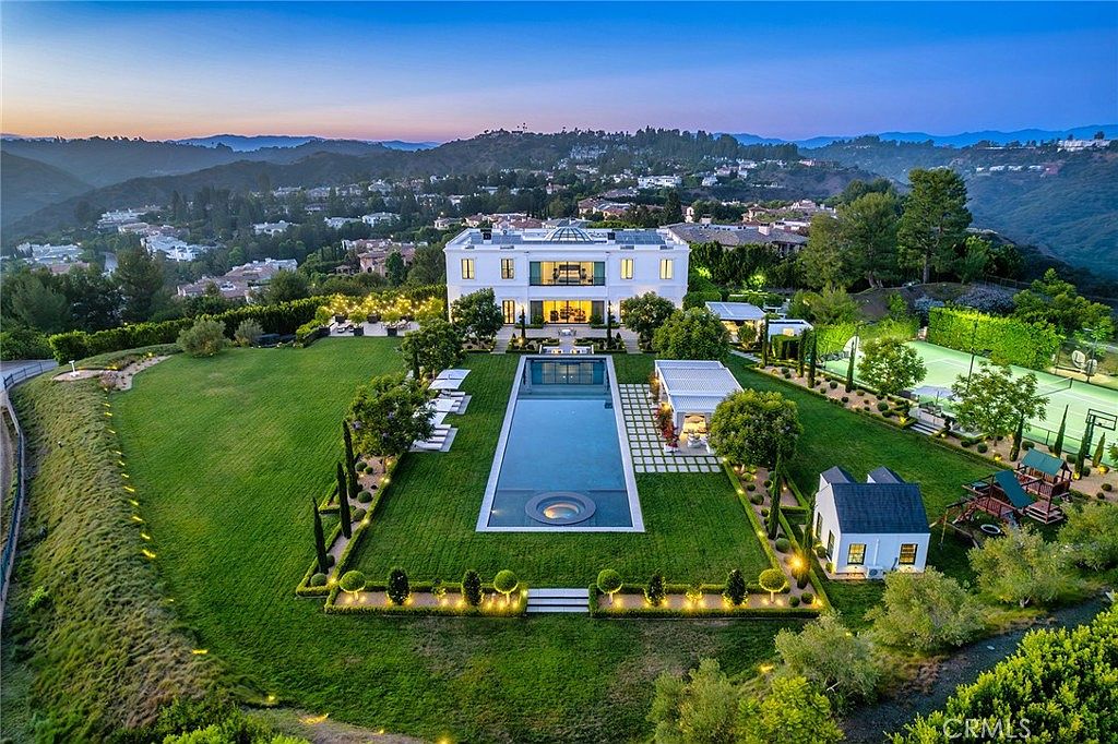 This aerial view showcases a luxurious estate with a sprawling green lawn, a rectangular swimming pool with a circular spa, and a grand white mansion as the focal point. The property also features a tennis court, a playhouse, and meticulously manicured landscaping, all set against a backdrop of rolling hills and a twilight sky. The overall impression is one of opulence and tranquility.