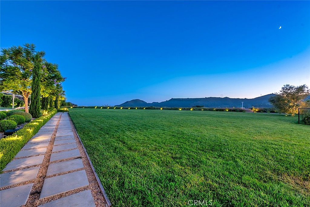 This image showcases a meticulously maintained yard or garden, featuring a lush green lawn that extends towards a distant mountain range under a clear blue sky. A stone pathway lined with trees and manicured shrubbery adds a touch of elegance and structure to the landscape. The scene evokes a sense of tranquility and spaciousness, highlighting the property's outdoor appeal.