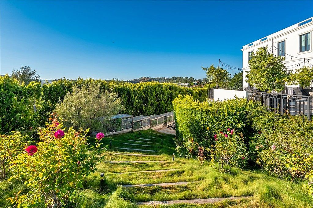 This image showcases a beautifully landscaped yard with a tiered garden featuring stone steps leading through lush greenery. The garden is adorned with flowering bushes and mature hedges, creating a serene and private outdoor space. A modern white building with a balcony overlooks the garden, adding a touch of architectural elegance.