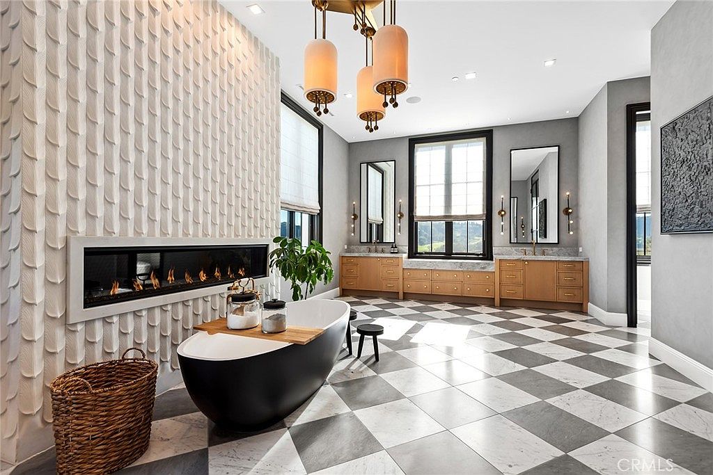 This is a luxurious primary bathroom featuring a freestanding black bathtub with a wooden tray, a modern fireplace with a textured white wall, and a checkered marble floor. The room is illuminated by stylish pendant lighting and natural light from the windows, creating a spa-like atmosphere. The perspective is from the doorway, showcasing the depth and elegance of the space.