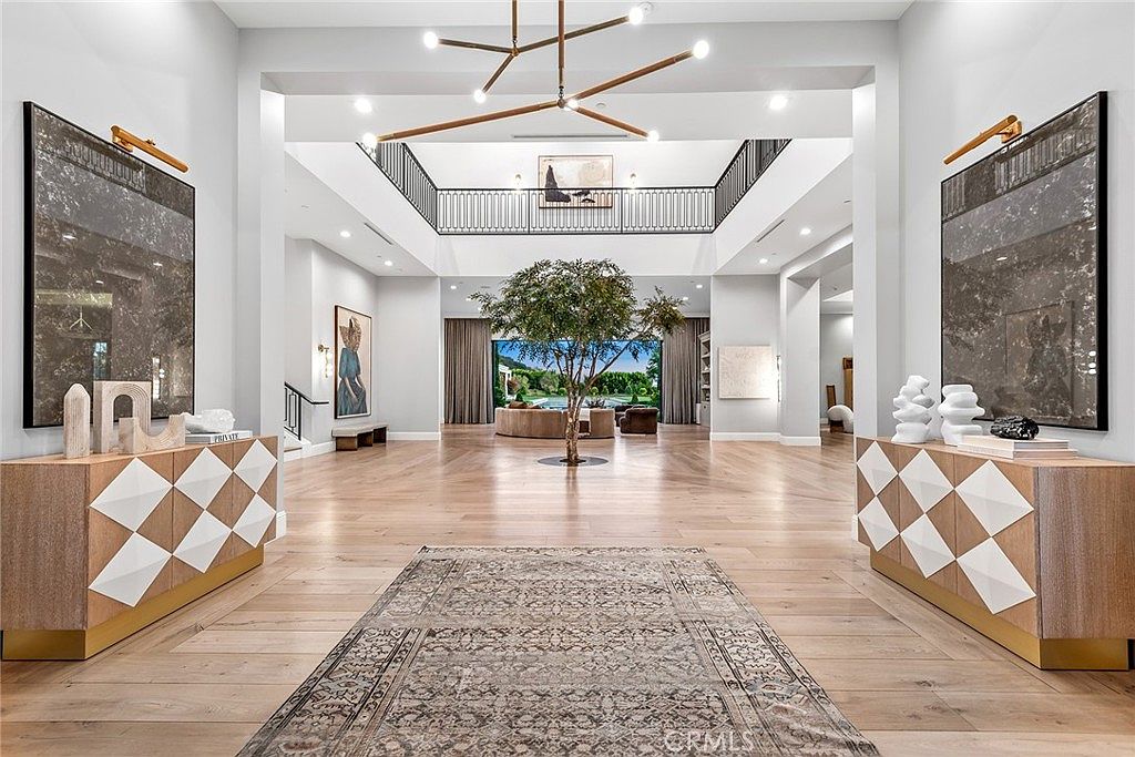 This grand foyer features soaring ceilings, light hardwood floors, and a striking central tree. Two decorative consoles flank the entrance, topped with artwork and sculptures. A large rug leads the eye towards a wall of windows overlooking a lush outdoor space, creating a seamless indoor-outdoor transition.