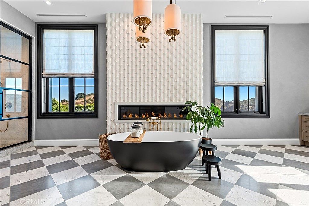 This is a luxurious primary bathroom featuring a freestanding black bathtub as the focal point, complemented by a modern fireplace and decorative wall paneling. The room is illuminated by stylish pendant lights and natural light from two large windows with black frames, offering views of the landscape. The checkered tile floor adds a touch of elegance, and a glass-enclosed shower is visible in the background, creating a spa-like atmosphere.