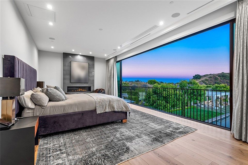 This is a luxurious primary bedroom featuring a large bed with a plush headboard and stylish bedding. A modern fireplace is set into a dark accent wall, and a large window offers a stunning view of the landscape and ocean. The room is spacious and well-lit, creating a serene and upscale atmosphere.