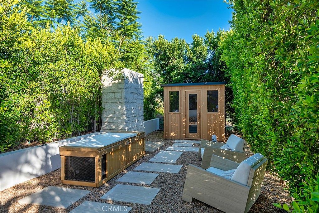 This image showcases a beautifully landscaped backyard featuring a sauna, a stone structure, and comfortable outdoor seating. The yard is surrounded by lush greenery, providing privacy and a serene atmosphere. Stone pavers create a pathway through the gravel, adding to the aesthetic appeal and functionality of the outdoor space.