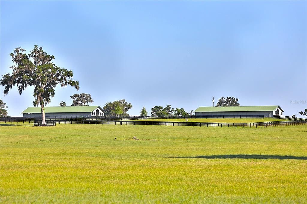 This expansive yard features a lush green lawn stretching towards two large, well-maintained buildings with green roofs, likely barns or stables. A dark wooden fence runs across the property, adding a touch of rustic charm. The clear blue sky and open space create a serene and inviting atmosphere, perfect for a rural estate.