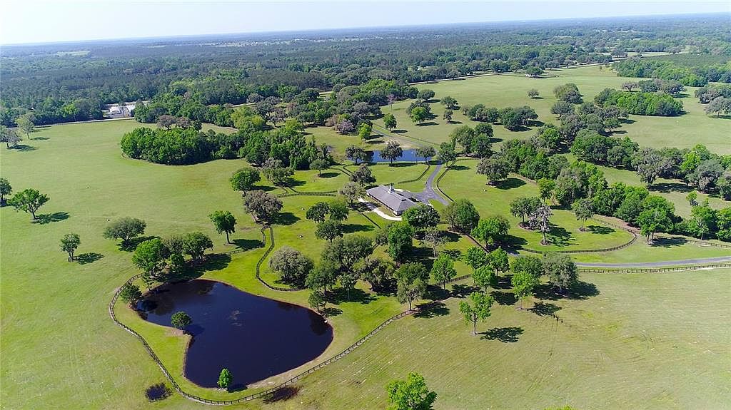 This aerial view showcases a sprawling estate with lush green pastures dotted with mature trees and two serene ponds. A well-maintained home is nestled among the trees, accessed by a winding driveway. The property is enclosed by fencing, creating a picturesque and private setting.