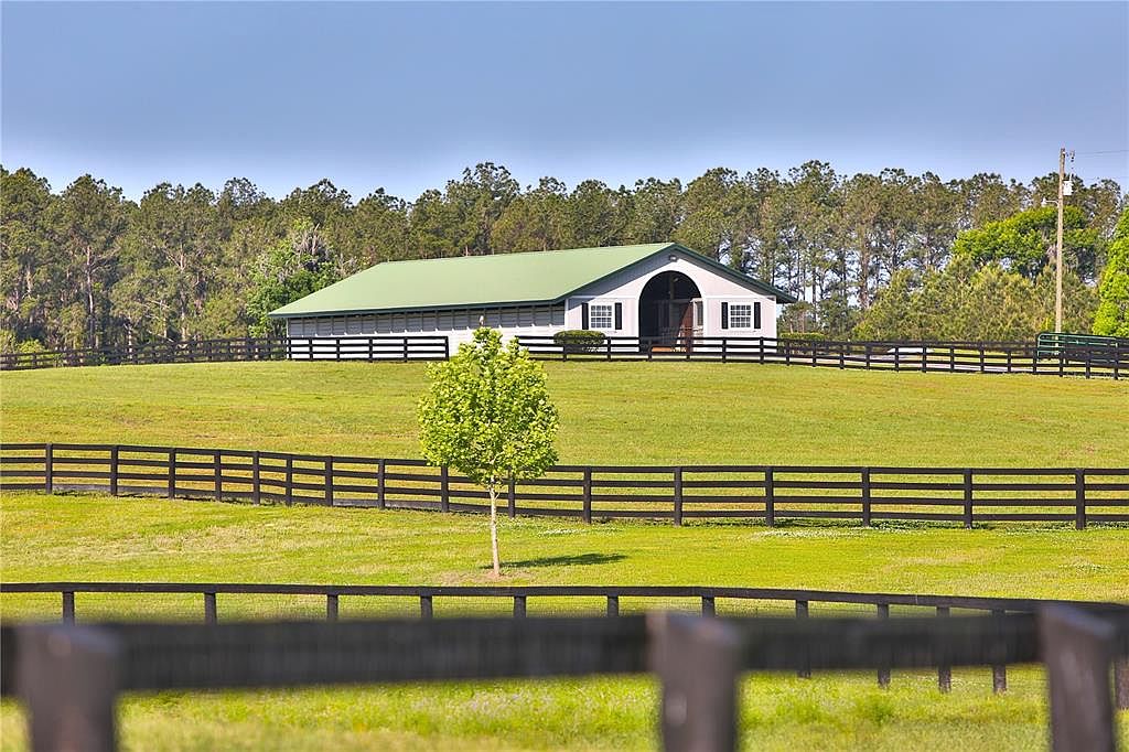 The image showcases a well-maintained yard with a large barn-like structure in the background. A dark wooden fence runs across the foreground and midground, adding a sense of enclosure and property definition. The lush green grass and mature trees create a serene and picturesque setting, ideal for a rural estate.